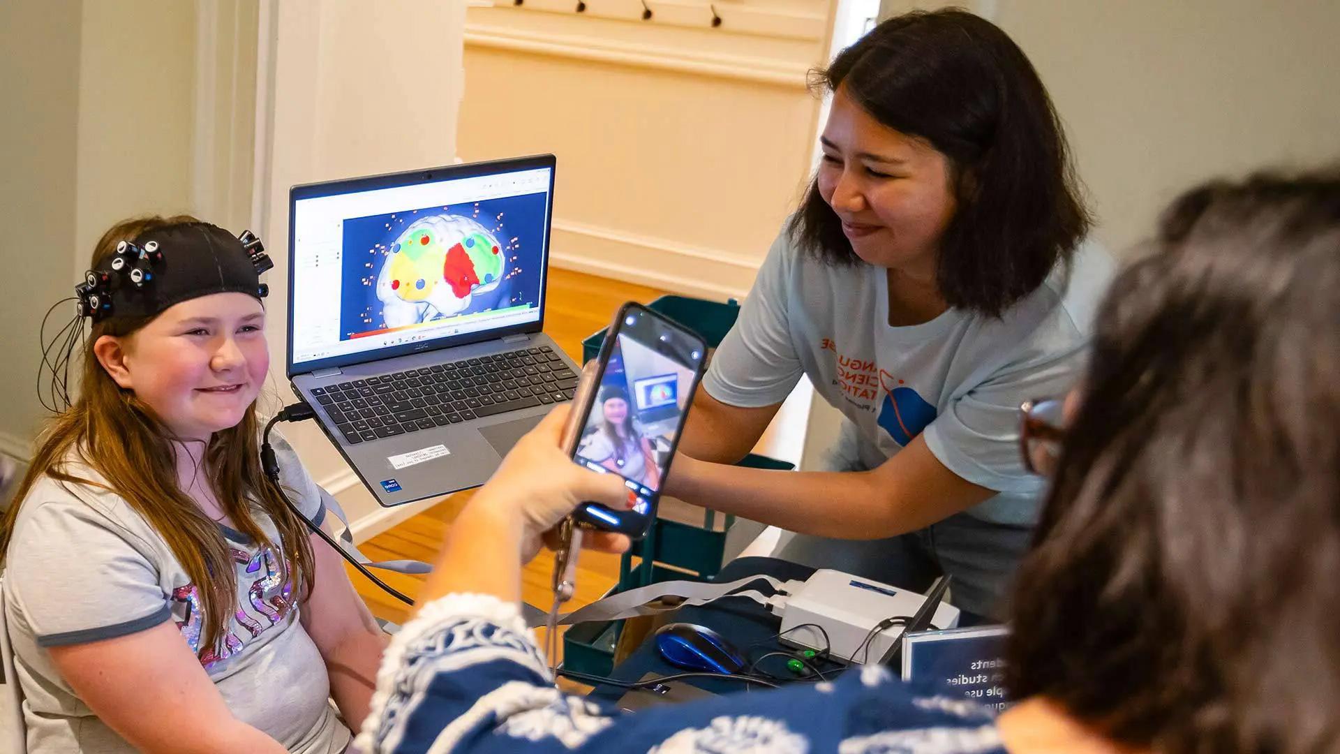 弗吉尼亚州亚历山大市的丽莎·奥多姆., 给她女儿拍了一张照片, 10岁的乔西, 因为她戴着一顶功能性近红外光谱(fNIRS)帽子, UMD人类发展博士生Gavkhar Abdurokhmonova拿着一台笔记本电脑，展示了她大脑中最活跃的部分.